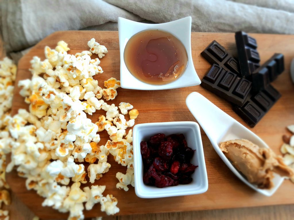 Popcorn, maple syrup, cranberries, dark chocolate, nut butter, oil, almonds on a chopping board